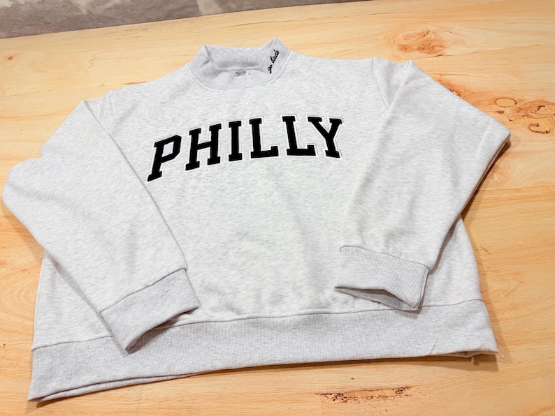 May include: A light gray heathered sweatshirt featuring the word PHILLY in bold black block letters across the chest. The garment has a mock neck collar with small black embroidered text on the right side, long sleeves, and ribbed cuffs.