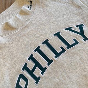 May include: A light gray heathered sweatshirt featuring a ribbed mock neck collar with the word birds embroidered in black script. The chest displays the word PHILLY in large, dark green block letters with a white outline, stitched onto the fabric.