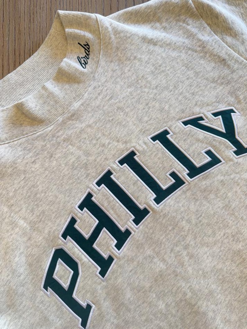 May include: A light gray heathered sweatshirt featuring a ribbed mock neck collar with the word birds embroidered in black script. The chest displays the word PHILLY in large, dark green block letters with a white outline, stitched onto the fabric.