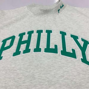 May include: A light gray heathered sweatshirt with a mock neck collar. The word PHILLY is embroidered in large green letters across the chest with a white outline. The text go birds is embroidered in green on the right side of the collar.