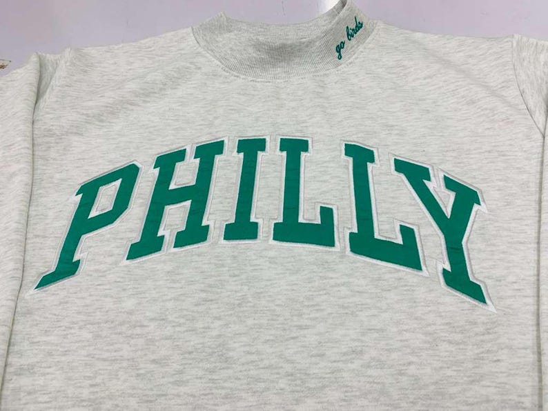 May include: A light gray heathered sweatshirt with a mock neck collar. The word PHILLY is embroidered in large green letters across the chest with a white outline. The text go birds is embroidered in green on the right side of the collar.