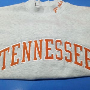 May include: A light gray sweatshirt with the word "TENNESSEE" in large orange letters across the chest. The sweatshirt has a mock neck collar with the words "go owls" embroidered in orange.