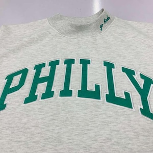 May include: A light gray heathered sweatshirt with a mock neck collar. The word PHILLY is embroidered in large green block letters across the chest. The text go birds is embroidered in green cursive on the right side of the mock neck collar.