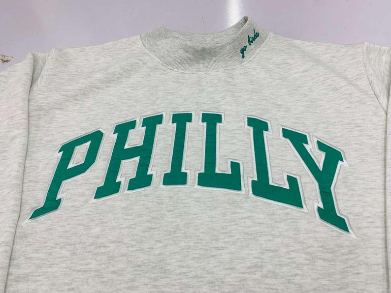 May include: A light gray heathered sweatshirt with a mock neck collar. The word PHILLY is embroidered in large green block letters across the chest. The text go birds is embroidered in green cursive on the right side of the mock neck collar.