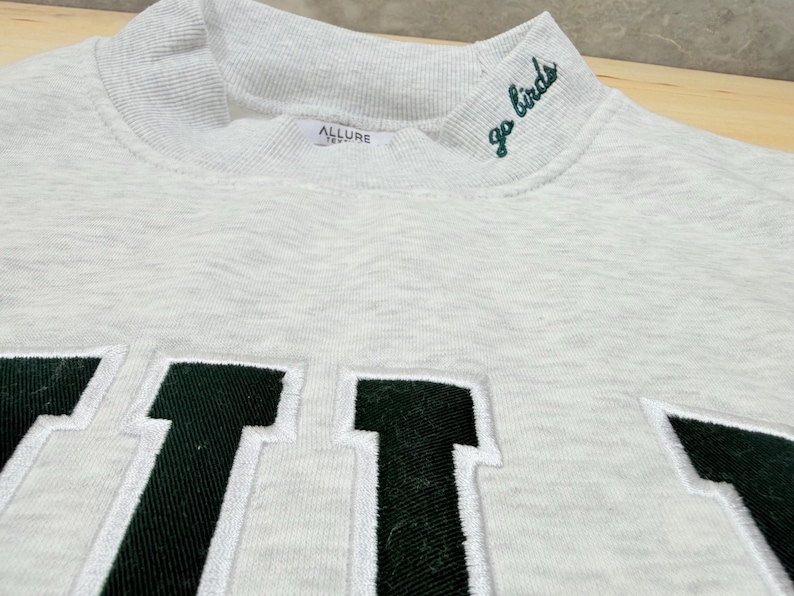 May include: A light gray heathered sweatshirt featuring a high mock neck collar with the embroidered text "go birds" in dark green. The front displays large, block-style letters in dark green with white stitching outlines. An Allure label is visible.