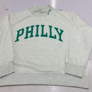 May include: A light gray heathered crewneck sweatshirt featuring the word PHILLY printed in bold green capital letters across the chest. The garment has long sleeves with ribbed cuffs, a ribbed hem, and a small green embroidered detail on the collar.