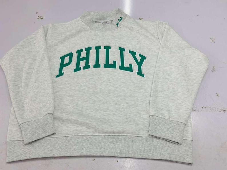 May include: A light gray heathered crewneck sweatshirt featuring the word PHILLY printed in bold green capital letters across the chest. The garment has long sleeves with ribbed cuffs, a ribbed hem, and a small green embroidered detail on the collar.