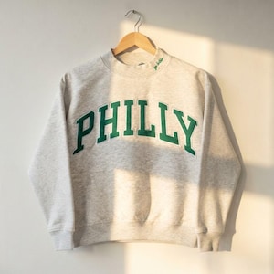 May include: A light gray heathered crewneck sweatshirt featuring the word PHILLY in large, dark green block letters across the chest. The neckline includes small green embroidered text that reads go birds, and the garment hangs on a wooden hanger.