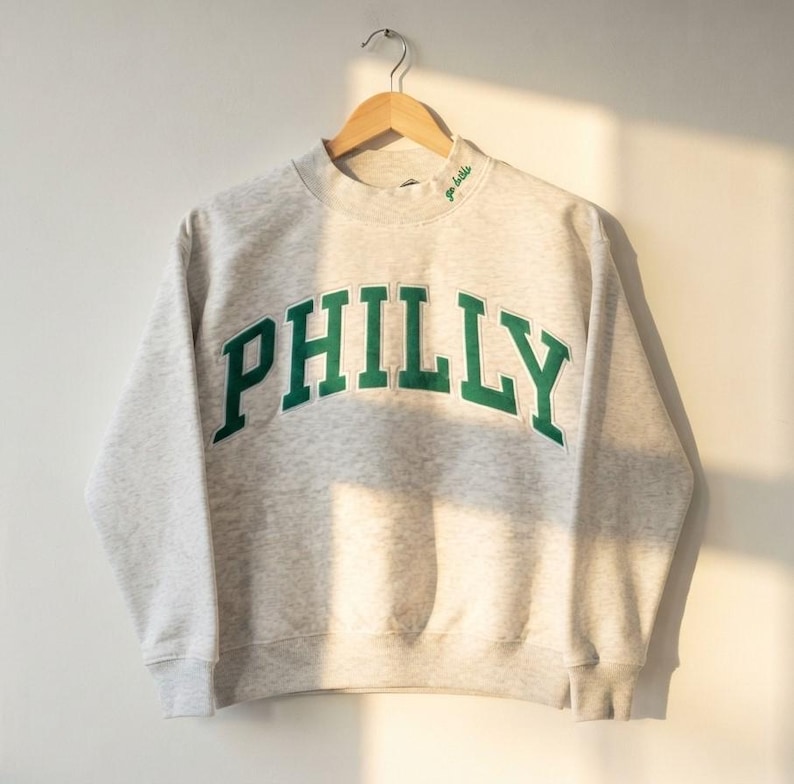 May include: A light gray heathered crewneck sweatshirt featuring the word PHILLY in large, dark green block letters across the chest. The neckline includes small green embroidered text that reads go birds, and the garment hangs on a wooden hanger.