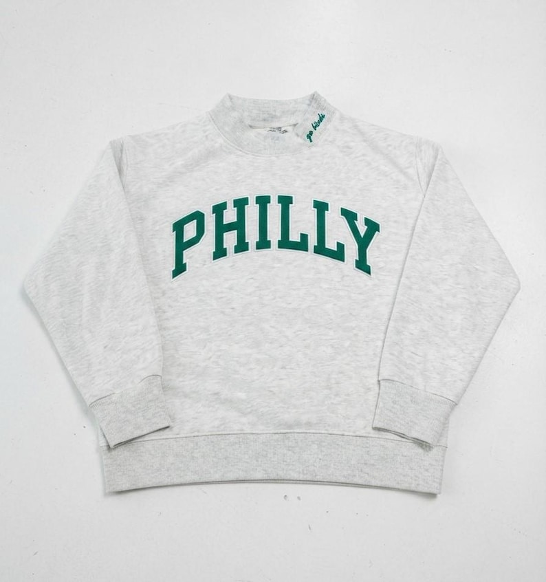 May include: A light gray heathered long-sleeve sweatshirt featuring the word PHILLY in large, dark green, arched block letters across the chest. The mock neck collar includes the small embroidered green text go birds on the left side.