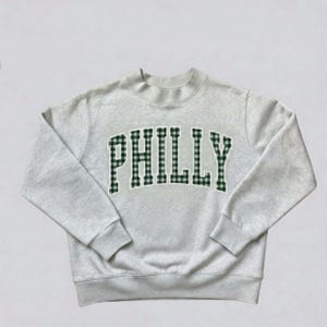 May include: A light gray crewneck sweatshirt featuring the word PHILLY in an arched, collegiate-style font. The letters are filled with a dark green and white gingham check pattern and outlined in a thin white border against the heathered fabric.