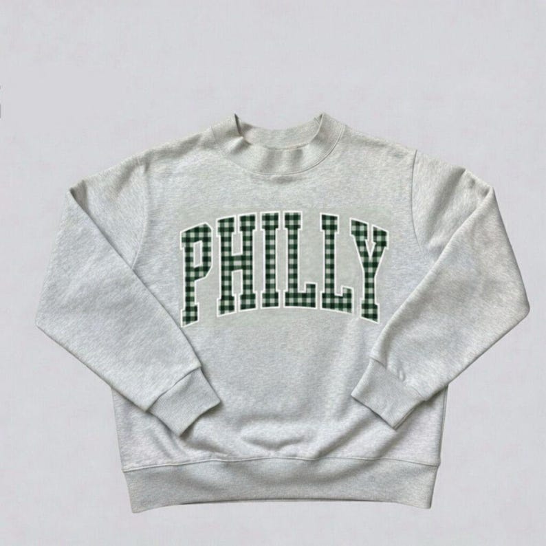 May include: A light gray crewneck sweatshirt featuring the word PHILLY in an arched, collegiate-style font. The letters are filled with a dark green and white gingham check pattern and outlined in a thin white border against the heathered fabric.