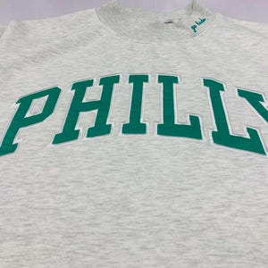 May include: A light gray heathered crewneck sweatshirt featuring the word PHILLY in large, bold green letters across the chest. The neckline includes a small embroidered green script text that reads go birds, positioned near the collar.