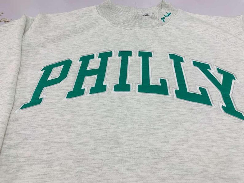 May include: A light gray heathered crewneck sweatshirt featuring the word PHILLY in large, bold green letters across the chest. The neckline includes a small embroidered green script text that reads go birds, positioned near the collar.