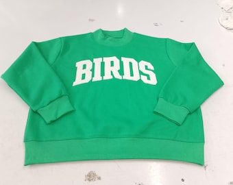 May include: A bright green long-sleeve sweatshirt featuring a mock neck collar and ribbed cuffs. The word BIRDS is printed in large, white, arched capital letters across the center of the chest. The garment has a straight hemline.