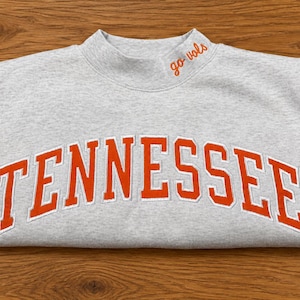 May include: A light gray mock neck sweatshirt featuring the word TENNESSEE in large, arched orange letters with white outlines across the chest. The collar includes the embroidered orange text go vols on the right side of the neckline.