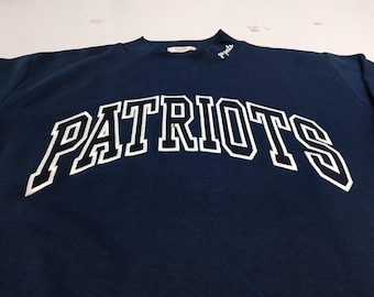May include: A dark navy blue long-sleeve sweatshirt featuring the word "PATRIOTS" in large, white, arched block lettering across the chest. The neckline includes a small white embroidered script text that reads "go pats" on the left side.