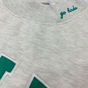 May include: A light gray heathered sweatshirt featuring a ribbed mock neck collar with the embroidered green text go birds. The garment includes large green block letters with white outlines stitched onto the front chest area of the fabric.