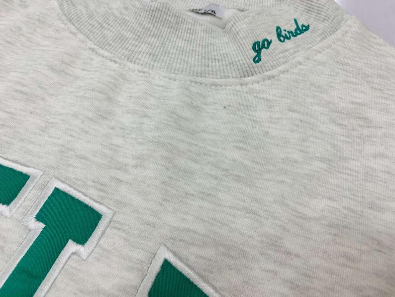 May include: A light gray heathered sweatshirt featuring a ribbed mock neck collar with the embroidered green text go birds. The garment includes large green block letters with white outlines stitched onto the front chest area of the fabric.