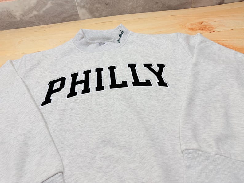 May include: A light gray heathered sweatshirt featuring the word PHILLY in large black block letters across the chest. The mock neck collar includes small green embroidered text that reads go birds, and the garment has long sleeves with ribbed cuffs.