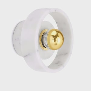 May include: A wall-mounted light fixture featuring a circular white marble frame with grey veining. The center contains a smaller circular marble base holding a polished gold spherical bulb cover that reflects the surrounding environment.