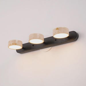 Peut inclure: Une applique murale composée d'une longue plaque de base horizontale noir mat. Trois abat-jours cylindriques à la texture beige tachetée rappelant la pierre sont fixés sur la base, chacun émettant une lumière blanche chaude et vive.