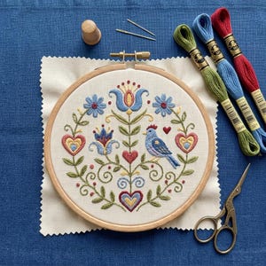 May include: A completed embroidery project in a wooden hoop, featuring a blue bird, hearts, and floral designs in blue, red, and green. Embroidery floss in green, blue, and red, along with scissors, a thimble, and needles are also visible.