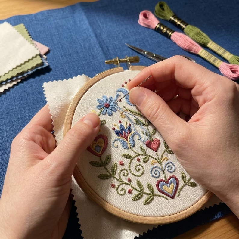 May include: An embroidery hoop with a floral design in progress. The design features blue, red, and green embroidered flowers and hearts. The needle is being threaded through the fabric. Embroidery floss and scissors are visible.