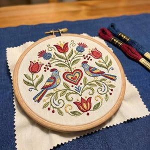 May include: A round, wooden embroidery hoop holding a detailed, colorful floral and bird design. The embroidery features red, blue, yellow, and green thread, with two birds facing each other above a heart. Two spools of thread are visible.