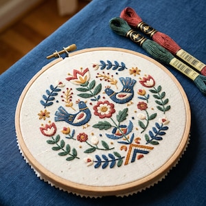 Op de afbeelding: Een ronde houten borduurring met een gebroken witte stof waarop een handgeborduurd ontwerp staat van twee blauwe vogels, omringd door rode, blauwe en groene bloemen- en bladmotieven. Twee strengen borduurgaren in rood en groen zijn inbegrepen.