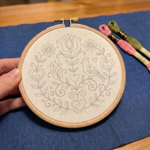 May include: A round embroidery hoop with a floral and bird design stitched in gray thread on a cream-colored fabric. The wooden hoop is held by a hand, with pink and green embroidery floss in the background. The design includes hearts, flowers, and a bird.