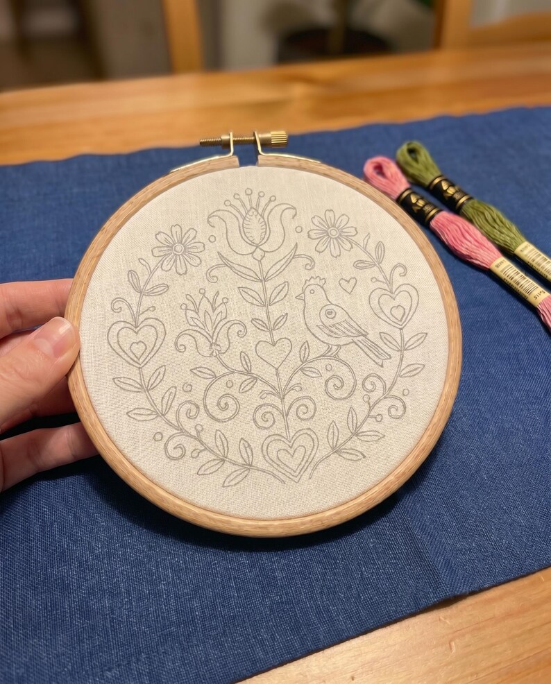 May include: A round embroidery hoop with a floral and bird design stitched in gray thread on a cream-colored fabric. The wooden hoop is held by a hand, with pink and green embroidery floss in the background. The design includes hearts, flowers, and a bird.