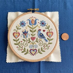 May include: A hand-stitched embroidery hoop featuring a folk art design. The design includes a blue bird, hearts, and floral elements in shades of blue, red, yellow, and green. The embroidery is set on a cream-colored fabric, framed by a wooden hoop.