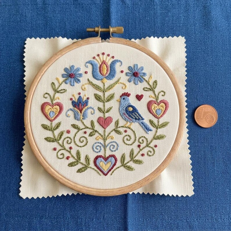 May include: A hand-stitched embroidery hoop featuring a folk art design. The design includes a blue bird, hearts, and floral elements in shades of blue, red, yellow, and green. The embroidery is set on a cream-colored fabric, framed by a wooden hoop.