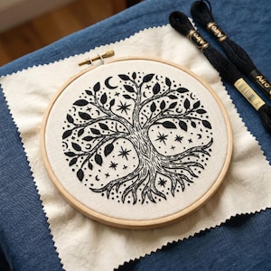 Può includere: Un ricamo in bianco e nero con un albero della vita in un telaio di legno. Il disegno include stelle, lune e foglie. Due rocchetti di filo da ricamo nero sono accanto al telaio su una superficie blu.