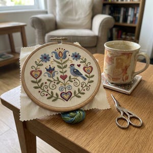 May include: A round embroidery hoop with a floral and bird design. The design features blue, red, and yellow flowers and hearts, with a blue bird in the center. The hoop is on a white fabric, with a cup of coffee and scissors nearby.