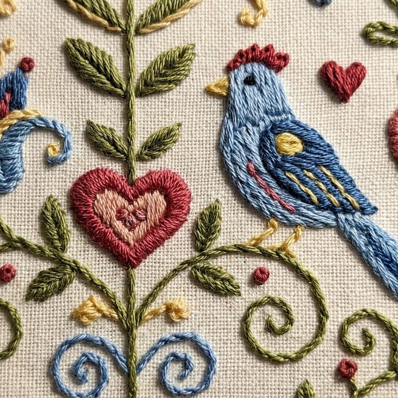 May include: Close-up of an embroidered design featuring a blue bird with a red crest, perched on a green vine. A red heart and a heart-shaped flower are also embroidered. The design is on a cream-colored fabric, with details in yellow and blue.