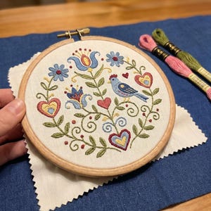 May include: A hand-stitched embroidery hoop featuring a blue bird, hearts, and floral designs in blue, red, and yellow. The embroidery is set in a wooden hoop, resting on a white fabric square, with spools of pink and green thread nearby.