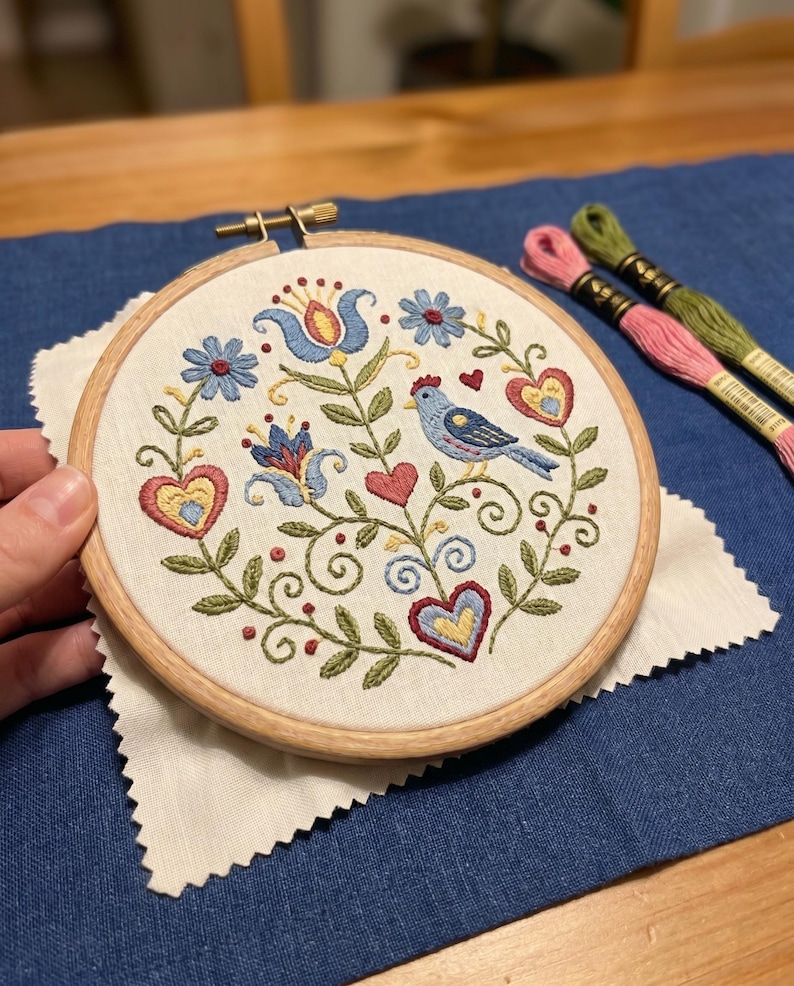 May include: A hand-stitched embroidery hoop featuring a blue bird, hearts, and floral designs in blue, red, and yellow. The embroidery is set in a wooden hoop, resting on a white fabric square, with spools of pink and green thread nearby.