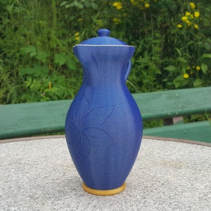 Puede incluir: Una jarra de cerámica azul con tapa y un diseño floral. La jarra tiene una base dorada y está sentada sobre una mesa gris.