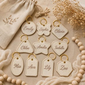 May include: A collection of ten beige fabric keychains in various shapes including circles, a butterfly, a square, a triangle, a star, a heart, ovals, rectangles, and an octagon, each featuring a gold metal ring and embroidered names.