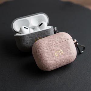 Peut inclure: Deux étuis personnalisés pour AirPod Pro, un gris et un rose, posés sur une surface sombre. L'étui gris est ouvert, révélant des écouteurs blancs. L'étui rose porte des initiales dorées. Un accessoire élégant.