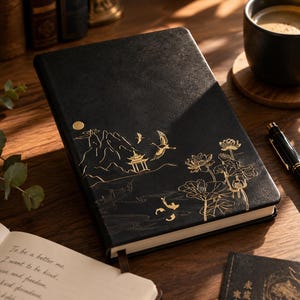 Peut inclure: Un carnet à couverture rigide noire orné d'illustrations dorées représentant un paysage de montagne, une pagode, une grue en vol et des fleurs de lotus. La couverture a une texture lisse et un marque-page ruban. Le carnet est fermé.
