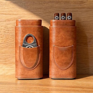 Può includere: Due astucci per sigari in pelle color cuoio con tasche cucite. L'astuccio a sinistra contiene un tagliasigari in metallo argentato. L'astuccio a destra contiene tre sigari con anelli circolari bianchi e neri visibili sulla parte superiore.