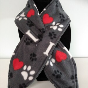 May include: Gray fleece scarf featuring a pattern of white paw prints, red hearts, and white bone shapes. The scarf is designed to be worn around the neck, offering warmth and a decorative touch. Ideal for pet lovers.