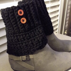 May include: A pair of gray suede ankle boots with black crochet boot cuffs. The cuffs are adorned with two orange buttons. The boots have a low heel and a buckle detail on the side.