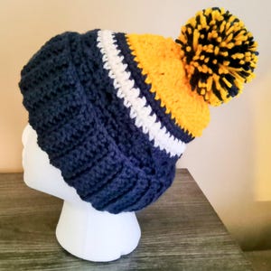 May include: A handmade, navy blue crochet beanie with a white and yellow stripe design. The beanie features a large, fluffy yellow and navy blue pom-pom on top. The hat is displayed on a white mannequin head.