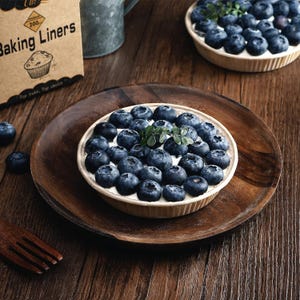 May include: A round paper baking liner filled with fresh blueberries and a small green sprig, resting on a dark wooden plate. A cardboard box labeled Baking Liners is visible in the corner, along with another filled liner in the background.