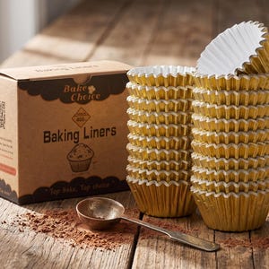 May include: A brown cardboard box labeled Bake Choice Baking Liners with a QR code and a muffin illustration, next to two stacks of metallic gold fluted paper baking liners and a small metal measuring spoon on a wooden surface.