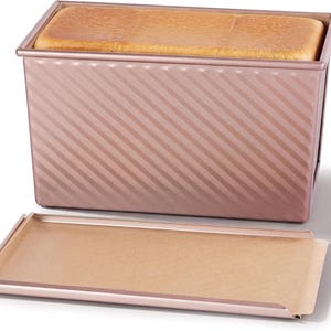 May include: A rectangular rose gold metal loaf pan with a diagonal ribbed texture on the sides. The pan contains a baked loaf of bread and is accompanied by a matching flat metal lid that is placed horizontally in front of the pan.